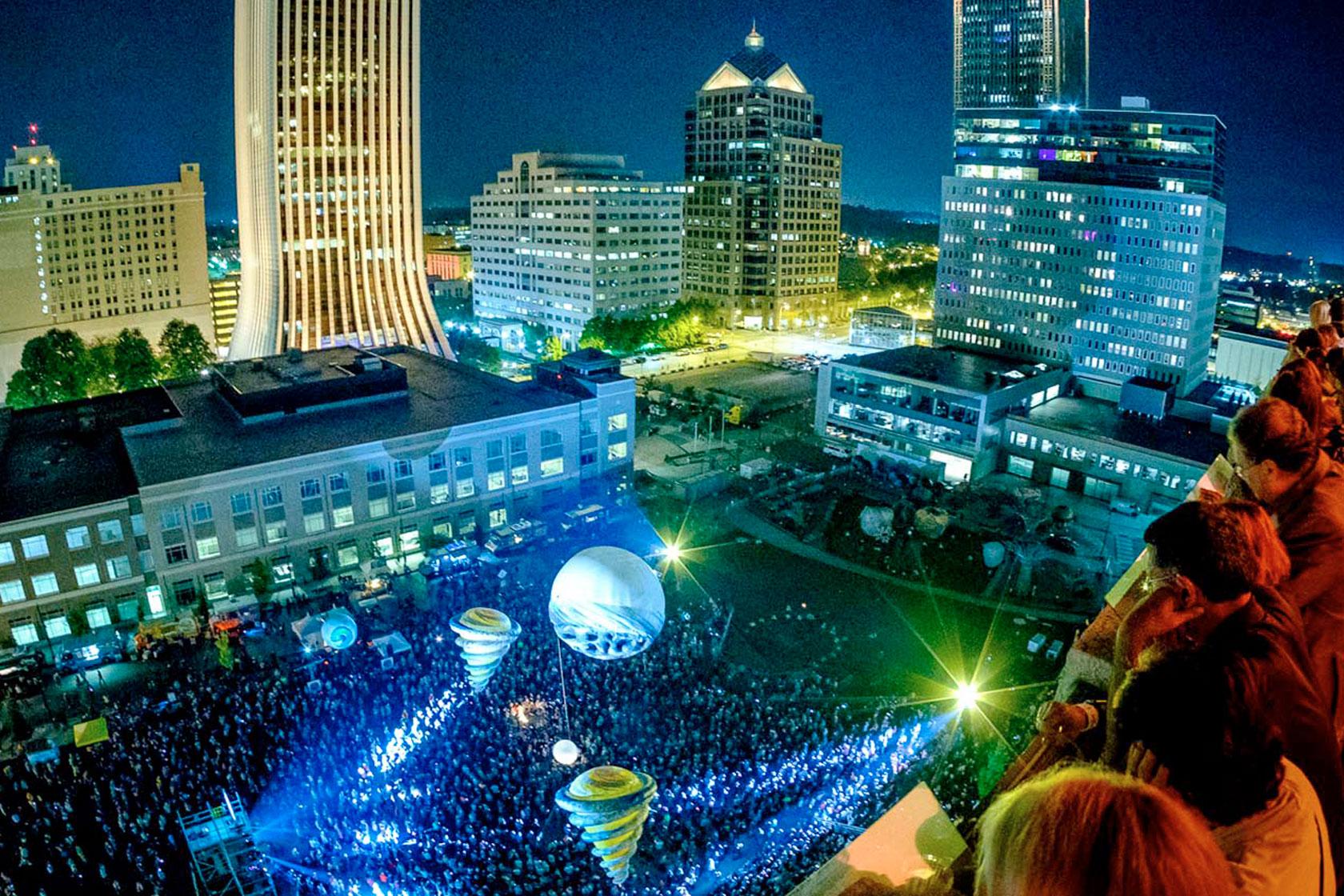 aerial view of large balloons flying over the crowd in downtown Rochester with the city skyline lit up in the background.